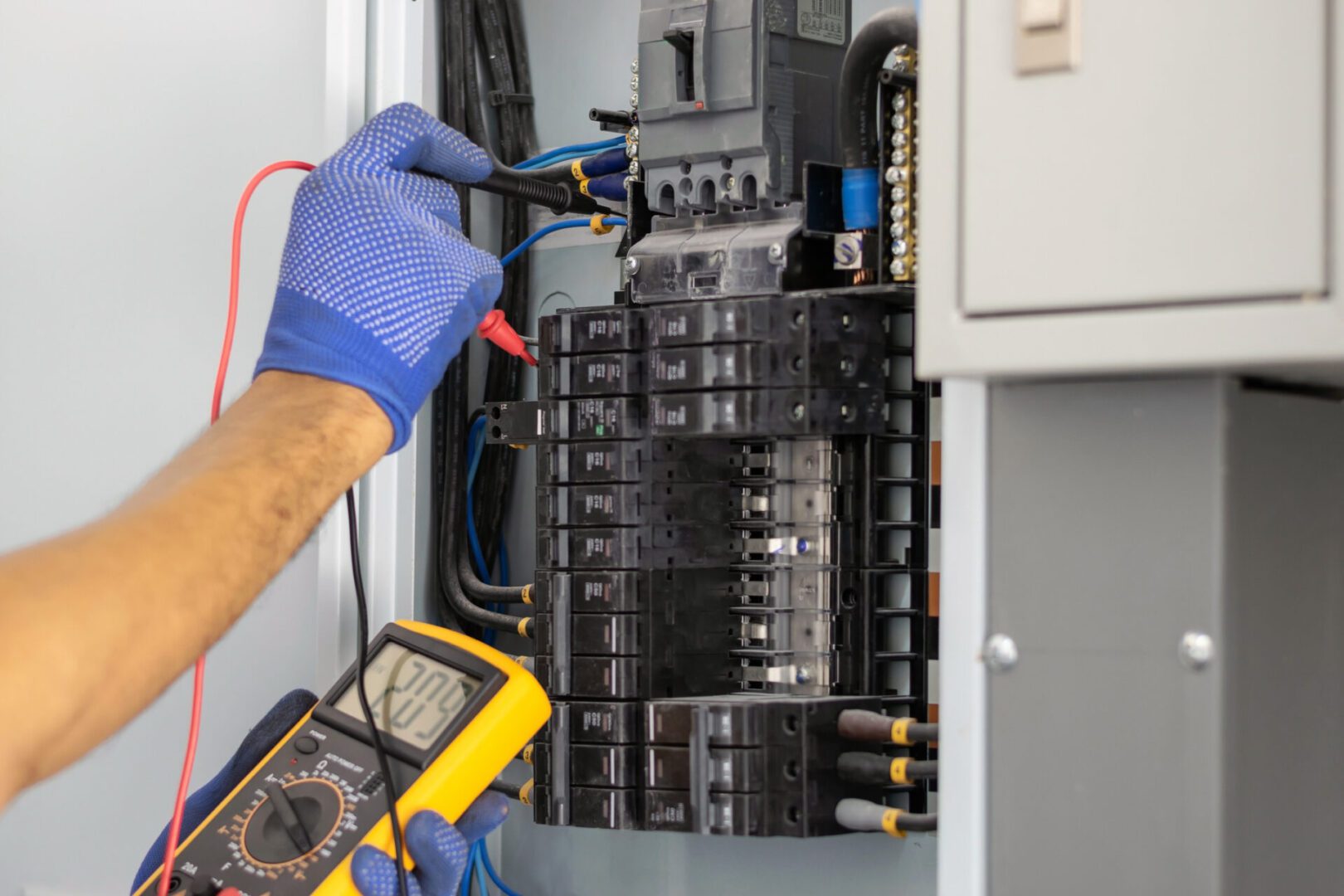 An electrician wearing blue gloves uses a multimeter to test an electrical panel, with wires connected for measuring voltage.