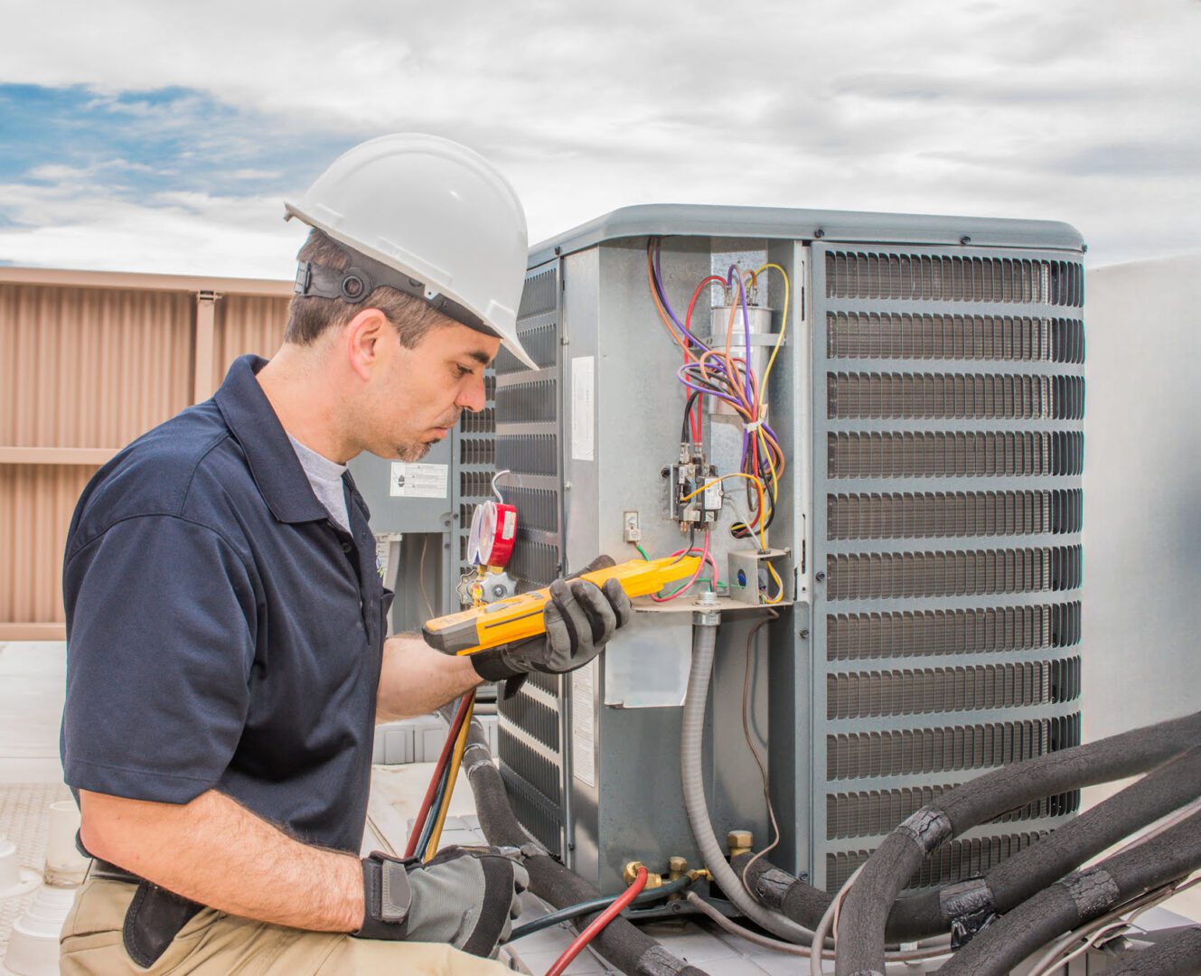 Technician in a hard hat uses a multimeter to test an HVAC unit on a rooftop.