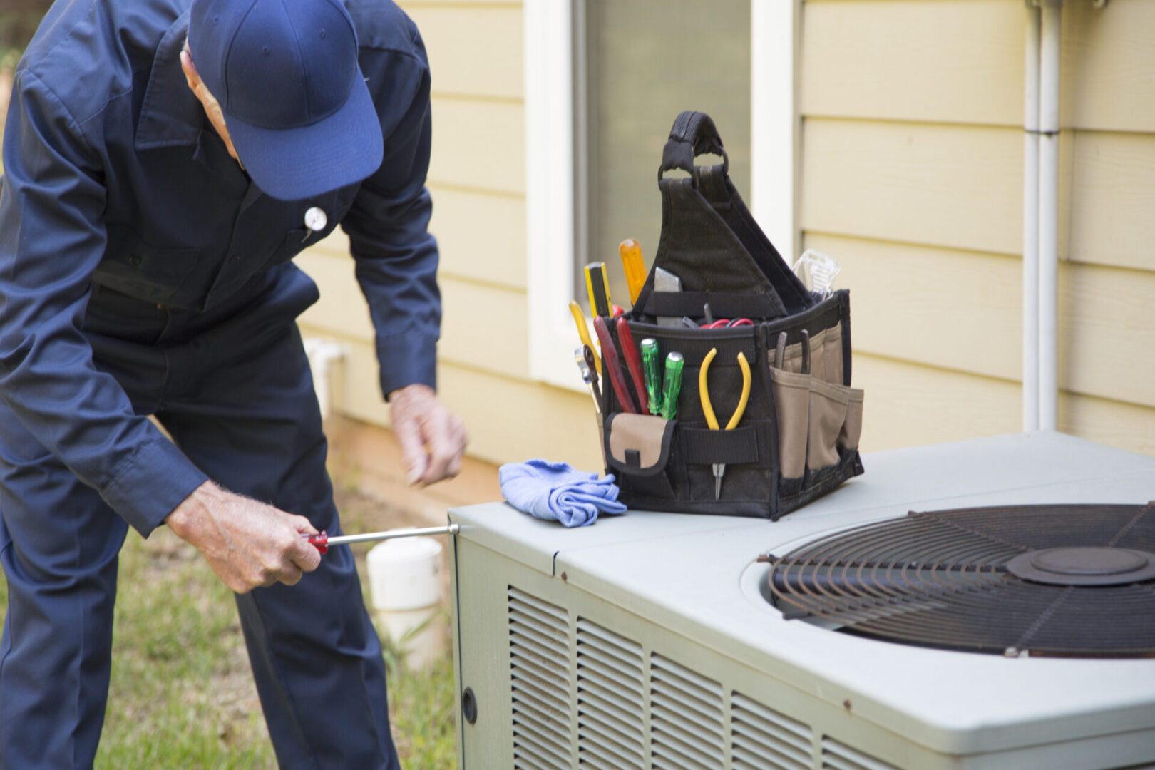 A technician in a uniform uses a screwdriver on an outdoor air conditioning unit, with a toolkit nearby.