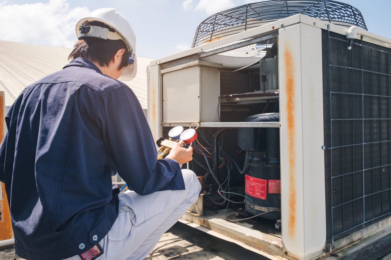 Technician in a blue shirt and helmet inspects HVAC system on a rooftop, holding gauges to check pressure levels.